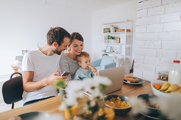 young family sitting at the kitchen island with their toddler son having lunch and using a laptop