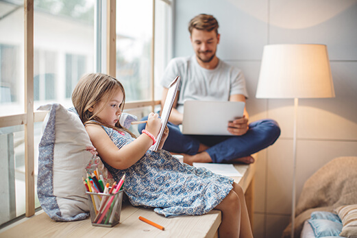 Dad using a laptop and daughter drawing on a notepad while sitting on a bench